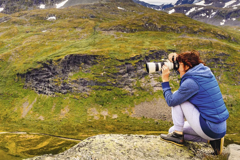 Mujer agachada sobre una roca fotografiando un valle de montaña con teleobjetivo, turismo de naturaleza que requiere tiempo y calma