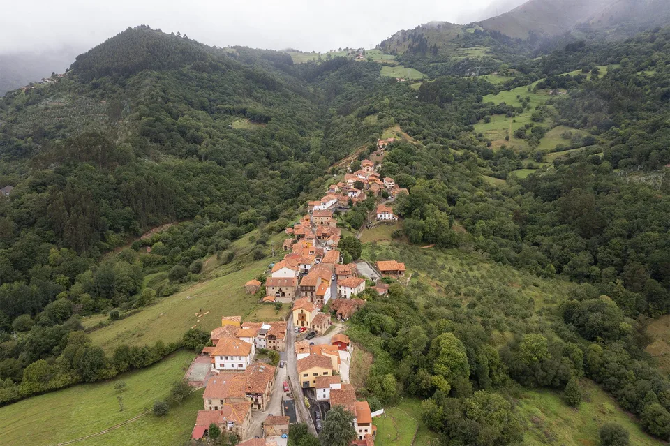 Vista aérea de un pueblo de tejados rojos en una ladera entre bosques y prados, destino de turismo rural para estancias largas